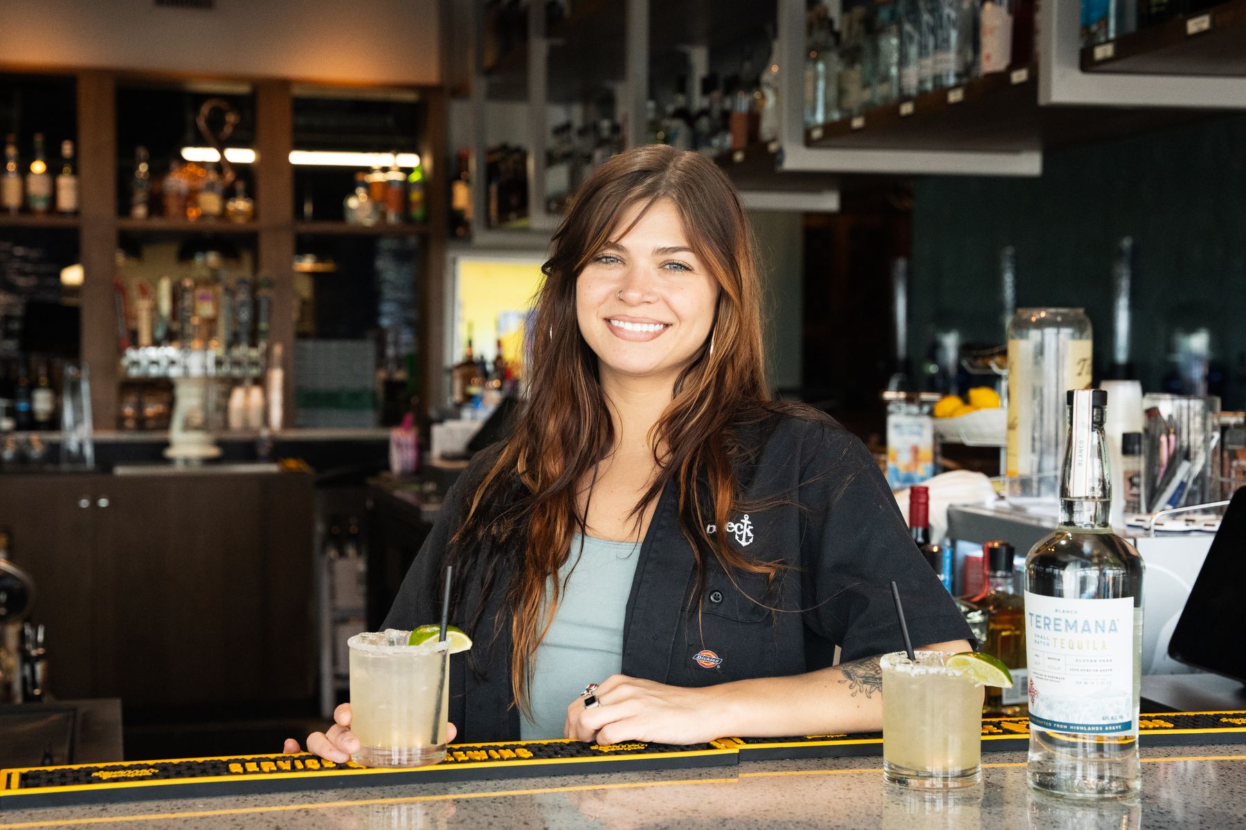 Smiling bartender leaning on the bar with two margaritas and a bottle of tequila.