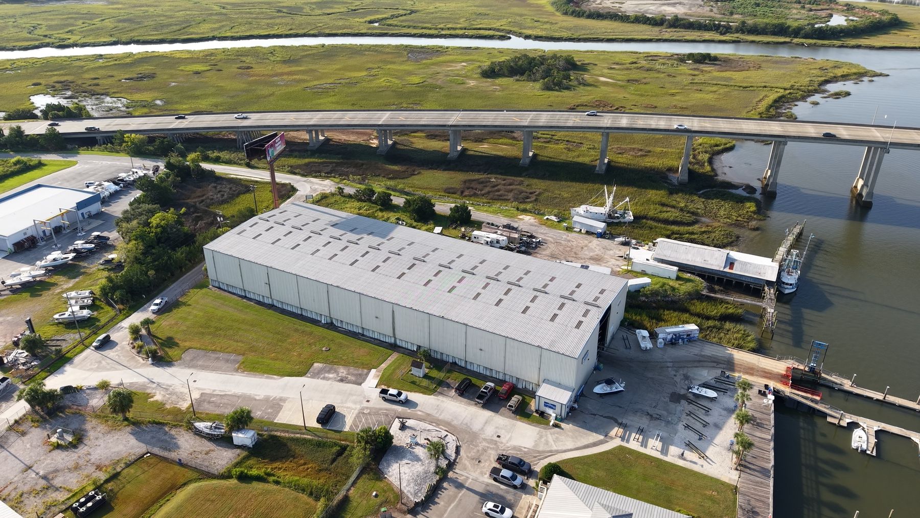 Aerial view of a waterfront boat storage building and marina beside a highway bridge and salt marsh.