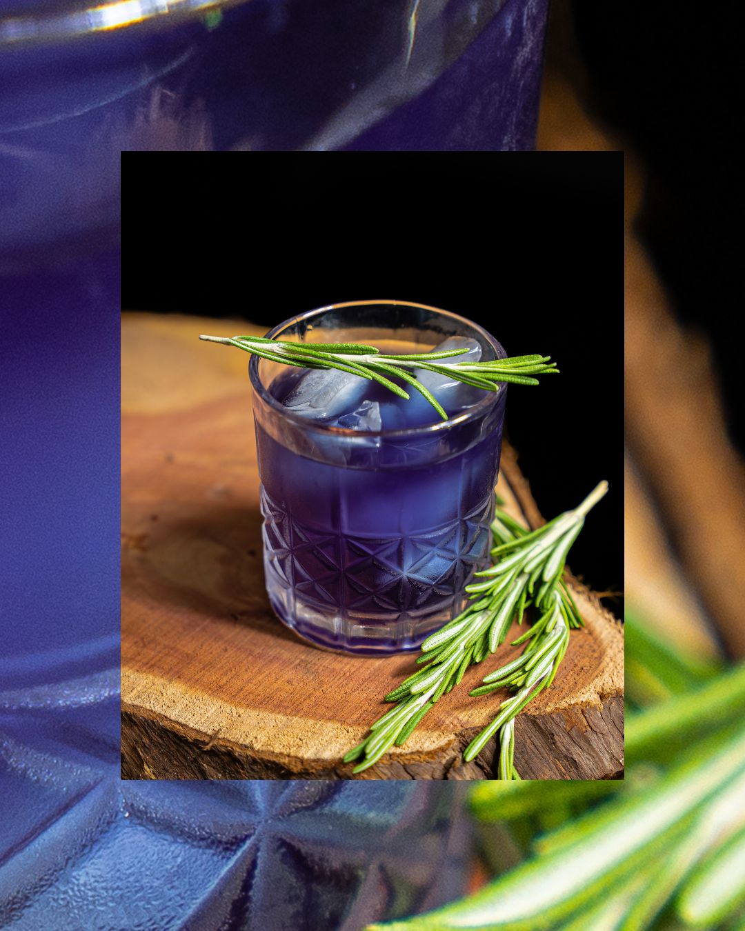 Purple cocktail garnished with fresh rosemary on a wooden board.
